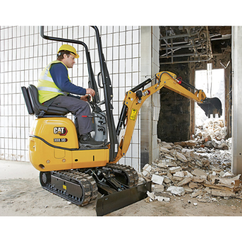 A construction worker operates a small yellow excavator demolishing an interior brick wall. The site is debris-laden, indicating ongoing construction.