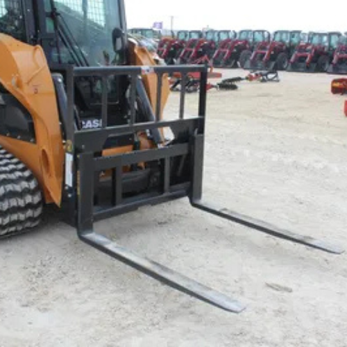 A skid steer loader with pallet fork attachment is parked on a dirt lot. In the background, several red tractors are lined up, creating an industrial setting.